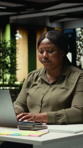 Vertical Video African american woman closing her laptop and office lamp, preparing to leave the building, going home at midnight. Employee working hard after hours, alone in dark space. Camera A.