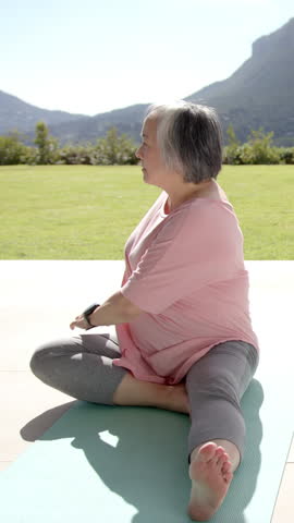 Vertical video: Sitting on teal mat Senior Asian woman stretching leg to side on terrace for yoga. Wellness, serenity, outdoor, relaxation, mindfulness, harmony, vertical video