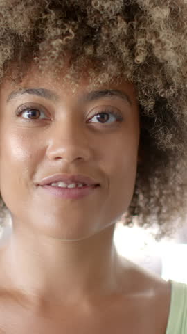 Vertical video: African American woman engaging camera shifting smile into laugh in bright room. Expressive, joyful, candid, vibrant, natural, emotion, vertical video