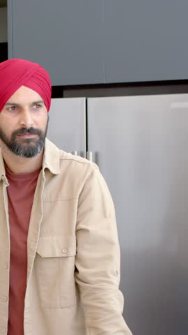 Vertical video: Indian man in red turban leaning at kitchen counter hearing voice smiling, nodding. Candid, modern, hospitality, friendly, candidness, lifestyle, vertical video