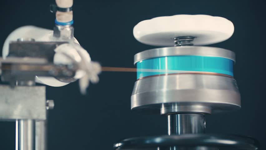 Slow-motion close-up of industrial coil winding machine in operation, type that winds flat copper wire. Detailed machinery spinning in a speaker factory.