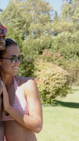 Diverse couple smiling in garden, wearing red and round black sunglasses and pink polka-dot bikini. Romance, outdoor, vibrant, nature, happiness, affection, lifestyle