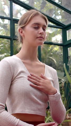 Vertical video: Woman placing hand on chest as sunlight filters in greenhouse, monitoring breathing. Meditation, mindfulness, serenity, relaxation, wellness, nature, vertical video