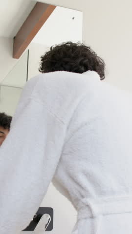 Man brushing teeth while checking smartphone in modern bathroom, with toothbrush and white bathrobe. Ardor, cleanliness, hygiene, minimalism, technology, self-care, wellness
