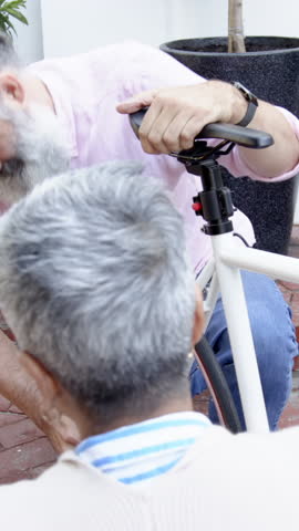 Senior friends repairing bicycle rear wheel on brick patio, with white frame and red-rimmed wheel. Collaboration, maintenance, craftsmanship, outdoor, rustic, teamwork, casual