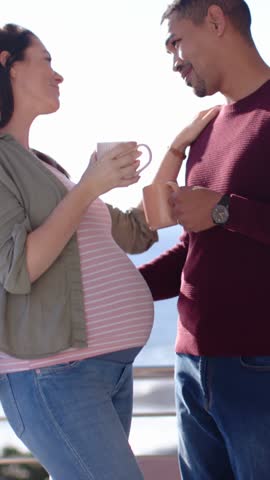 Standing diverse couple holding ceramic mugs on seaside balcony, with metal railing and water. Romance, serenity, happiness, leisure, lifestyle, coastal, pastel