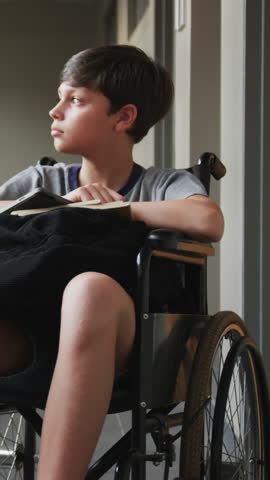 Child boy sitting in manual wheelchair in hallway, holding folded arms on notebook and smartphone. Disability, accessibility, inclusivity, youth, education, modern, comfortable