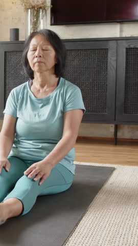 Vertical video: Senior Chinese woman starting meditation at home, alternating leg poses on yoga mat. Meditation, mindfulness, wellness, serenity, aging, tranquility, vertical video