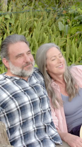 Vertical video: Senior couple sitting on bench in park, head leaning, man pointing, sun hat beside. Romantic, companionship, serenity, outdoor, leisure, affection, vertical video