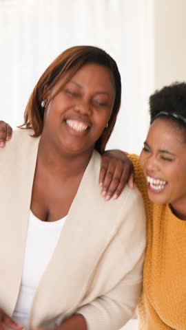 Vertical video: African American female friends clapping, laughing and hugging at home sharing joy. Friendship, joy, togetherness, casual, warmth, happiness, vertical video