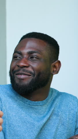 Vertical video: African American man smiling at offscreen friend, sipping mug on porch and laughing. Relaxation, friendliness, leisure, outdoor, casual, warmth, vertical video