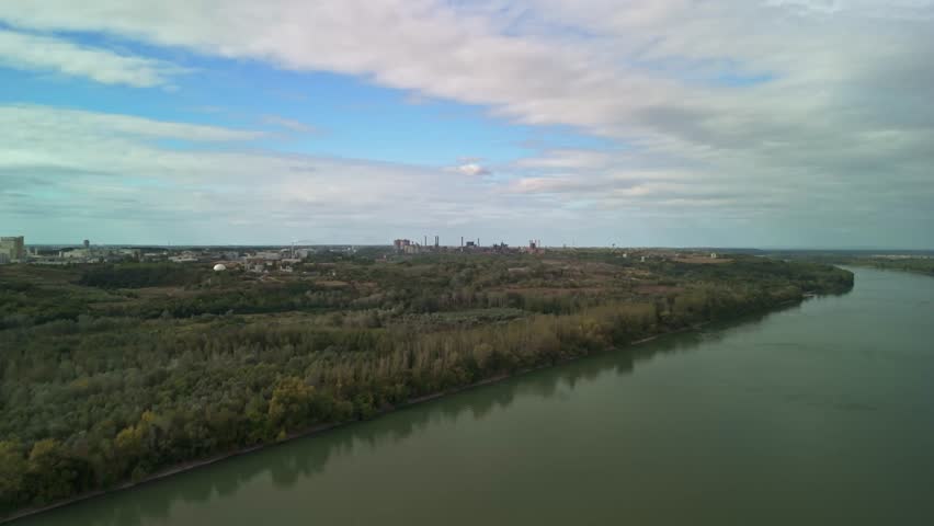 Aerial drone view of Dunaújváros, Hungary, showing the city’s industrial skyline and factory chimneys in the distance across the Danube River under partly cloudy skies.