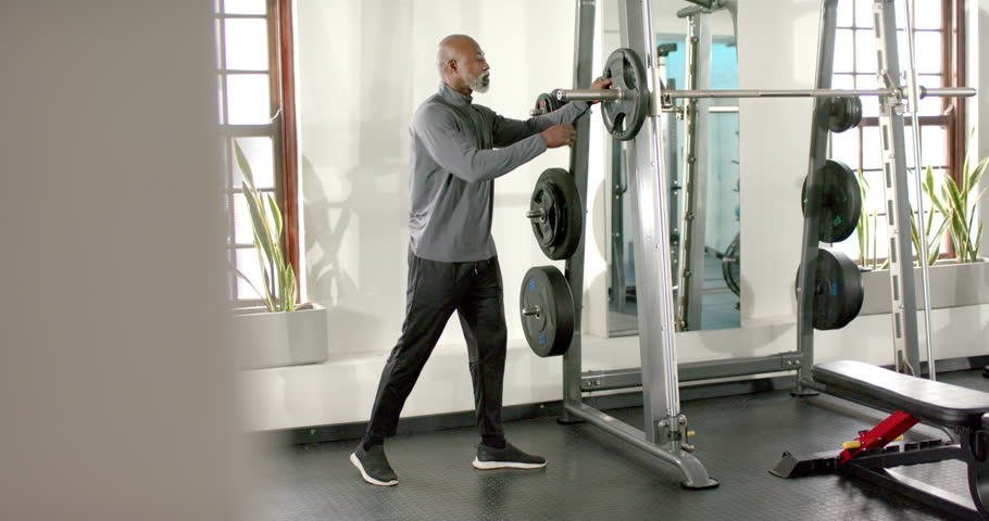 African American man approaching Smith machine, loading weight plates from rack, preparing workout. Fitness, strength, weightlifting, gym, workout, motivation, masculinity