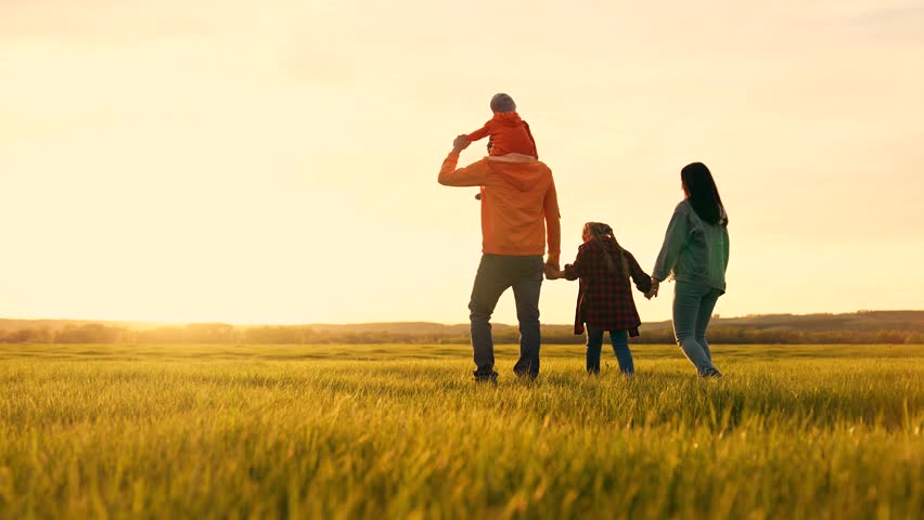 Family walking across green field toward setting sun with father carrying son on shoulder and mother holding daughter hand in tall grass at golden sunset family walk child play in rural landscape
