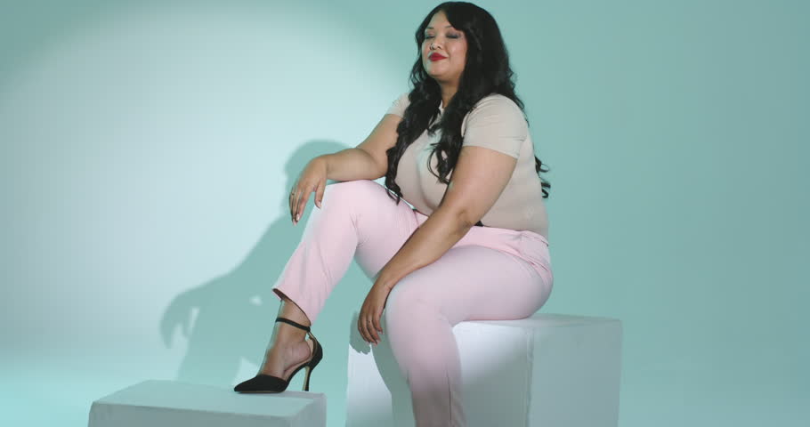 Upon camera rolling mid adult woman posing, curling fingers, laughing on white cubes in mint studio. Modern, elegance, pose, minimalism, fashion, lifestyle, studio