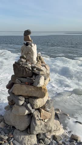 Vertical video of Baikal Lake on frosty December day. Close-up of stone pyramid of tourists on icy coast and white ice floes near water during freeze-up. Typical Baikal lakeside landscape. Winter tour