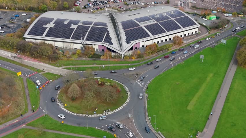 Drone flies towards Harlow Leisurezone, Essex. The modern angular building has two connected wings forming a 45° bend. Its south-facing roof is lined with solar panels by a main road and roundabout.