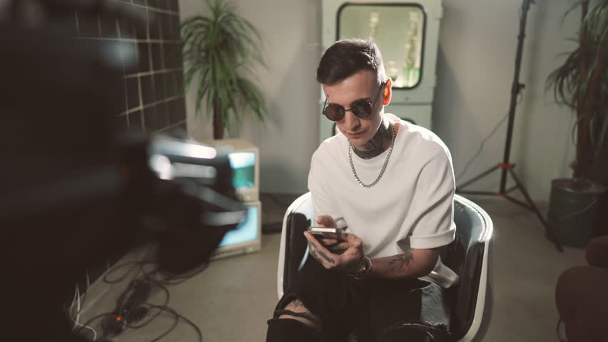 Trendy blogger or rock star recording vlog for social media, portrait in studio. Young adult male person with tattoos, cool sunglasses and accessories posing for camera, professional shooting indoors