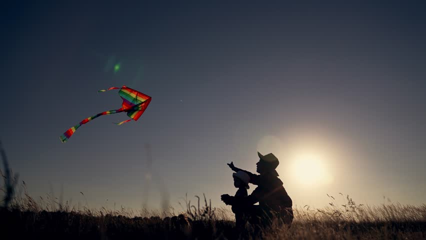 Flying kite with parent and child silhouette on grassy field at sunset with colorful kite soaring in open sky while parent and child point and play with family close to tall grass under warm soft sun