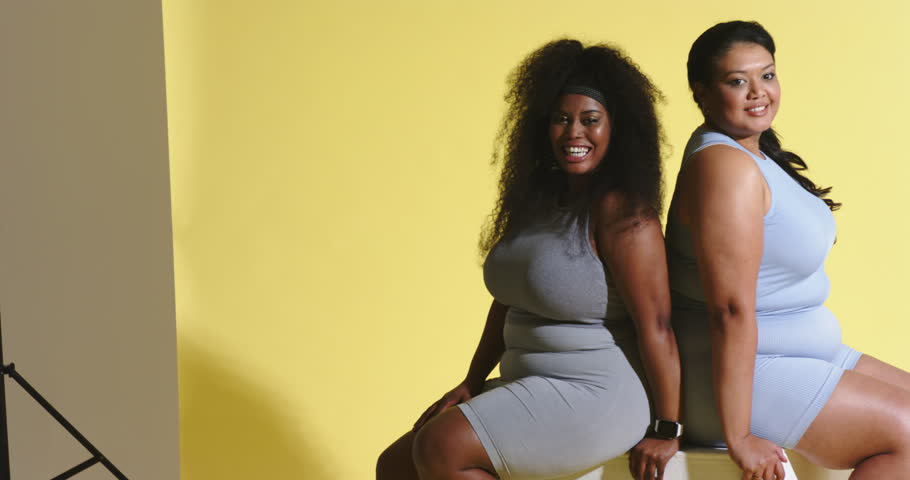On cue Diverse female models shifting on stools, leaning and laughing against yellow backdrop. Friendship, camaraderie, playful, cheerful, dynamic, studio, portrait