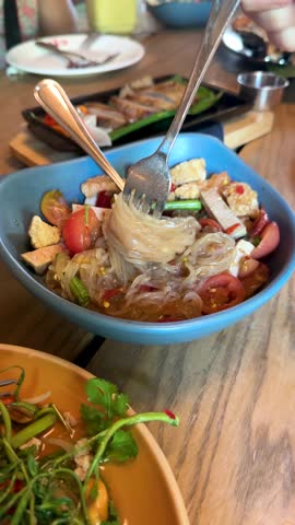 Hand lifts glass noodles from vibrant Thai salad in bright, casual Bangkok restaurant setting.