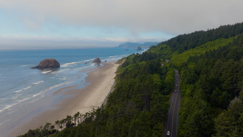 Cinematic aerial slow push along Douglas firs and the coastline road at Arcadia Beach in Oregon, moving toward Cannon Beach with sea stacks and Haystack Rock under a pretty partly cloudy sky.