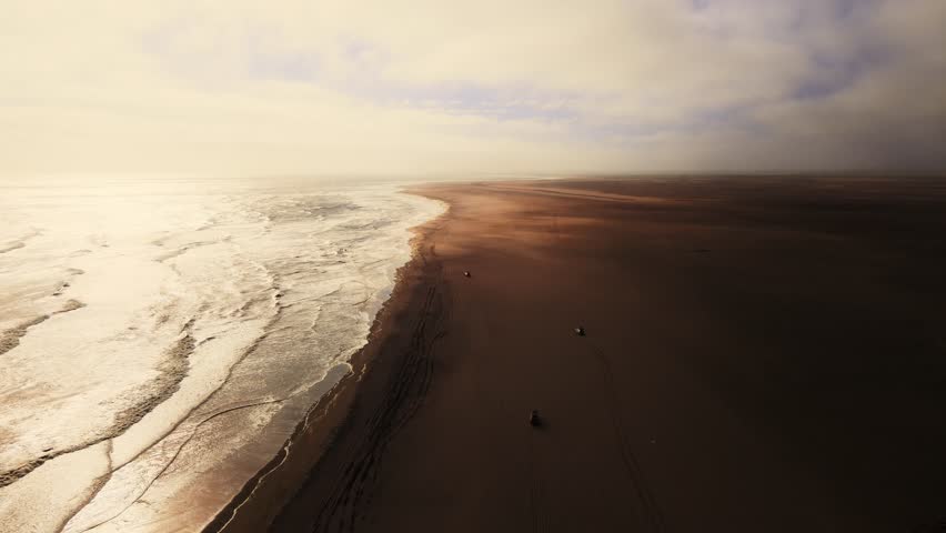 Several jeeps drive along the edge of the Namib Desert beside the Atlantic Ocean at sunset, filmed by drone, where golden dunes meet the blue waves, creating a stunning contrast of land and sea.