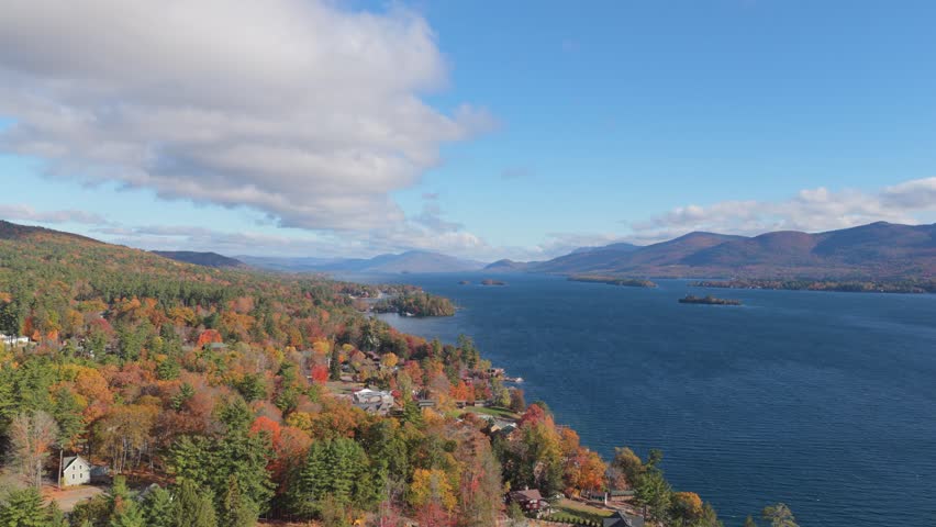 6K aerial view of Lake George surrounded by vibrant fall foliage, blue water, and distant mountains under a bright sky in upstate New York.