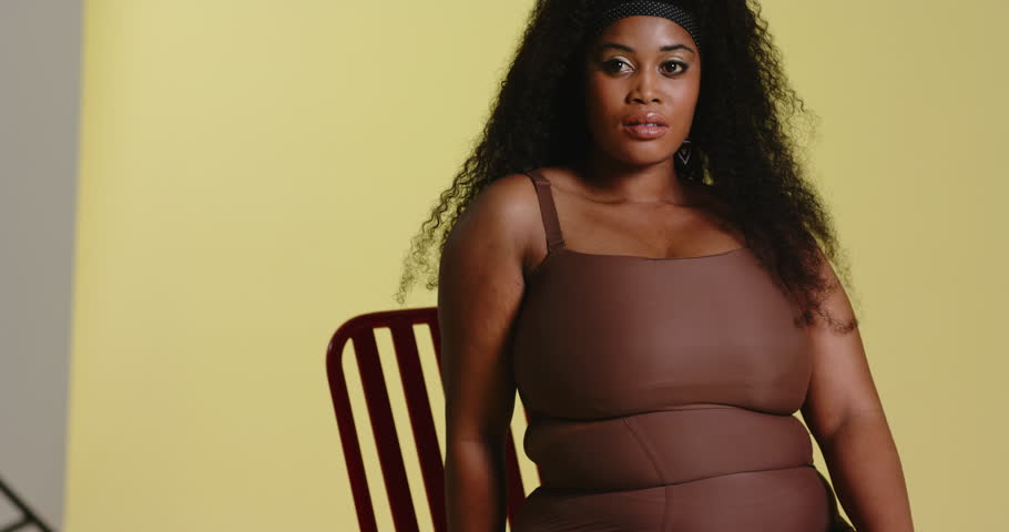 At start African American woman shifting weight, turning head left on chair before yellow backdrop. Portrait, wellness, serenity, minimalist, contemplative, elegance, beauty