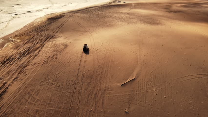 Several jeeps drive along the edge of the Namib Desert beside the Atlantic Ocean at sunset, filmed by drone, where golden dunes meet the blue waves, creating a stunning contrast of land and sea.