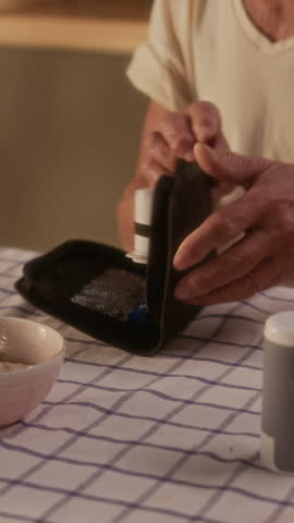 Vertical medium close-up of elderly woman in glasses sitting at table in kitchen after breakfast, unwrapping diabetes kit, preparing to take finger stick glucose test