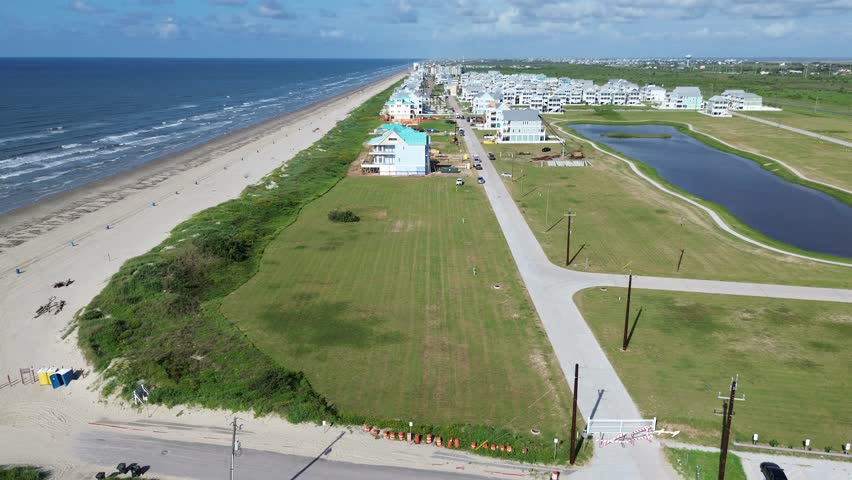 Galveston Beachside Village near Sunny Beach shows long sandy shoreline with blue trash bins, grassy buffer zones, and pond surrounded by coastal homes and manicured lawns, Texas vacation trips. USA