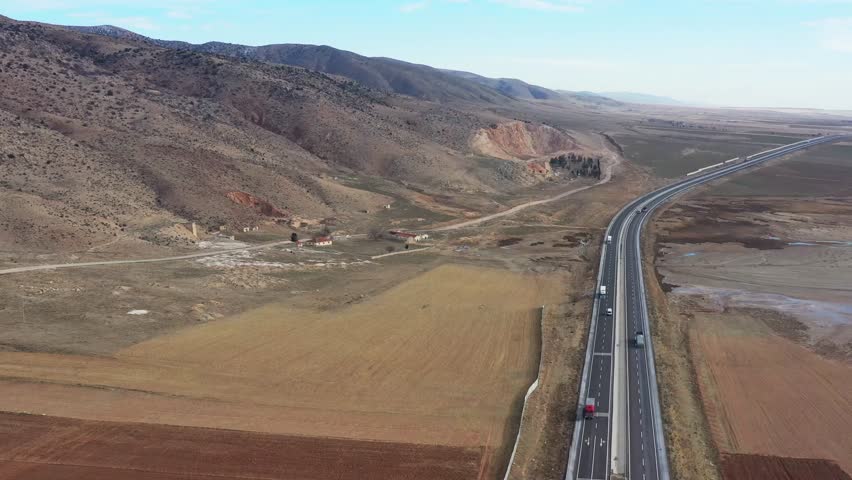 Aerial view of a long highway cutting through arid landscapes and rolling hills under a clear sky, near Tuz, Ankara, Turkey.