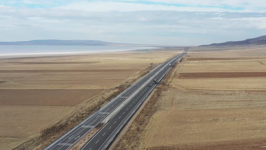 Aerial view of a long highway cutting through vast fields towards the white expanse of Tuz Lake under a cloudy sky, Tuz, Ankara, Turkiye.