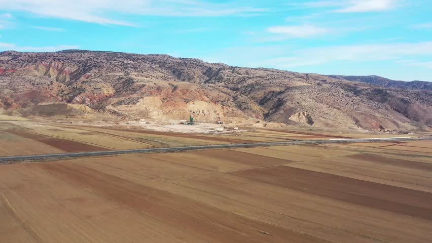 Aerial view of harvested fields contrasting with the rugged mountains and a road with moving vehicles, Tuz, Ankara, Turkiye.