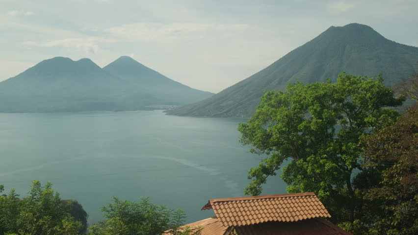Aerial drone footage gliding over the calm waters of Lake Atitlán, Guatemala, surrounded by majestic volcanoes in soft morning light. The movement captures the essence of serenity