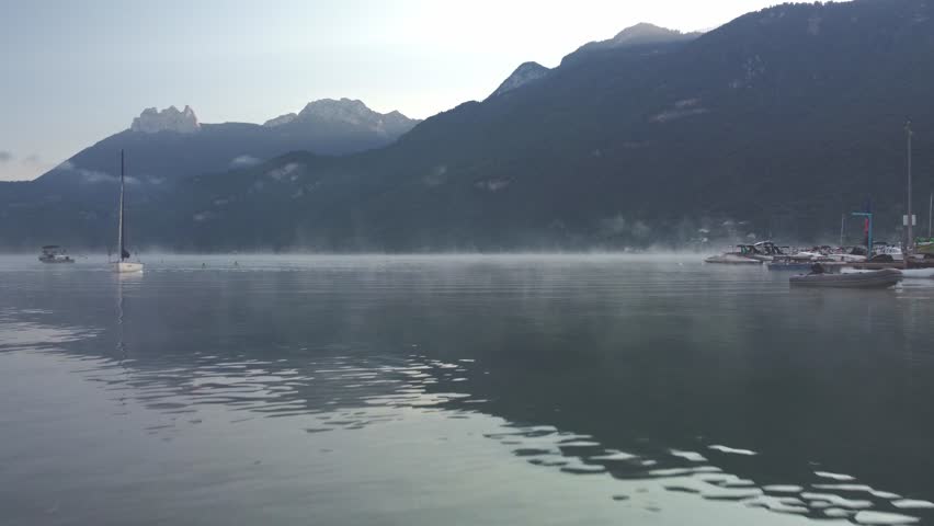 Low flying drone footage over the water of Lake Annecy on a misty morning. Some boats moored at a marina can be seen initially. Mist steams off the water as the sun rises over the mountains. France.