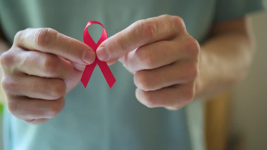 Close-up shot of male hand holding red satin ribbon symbolizing HIV AIDS support remembrance and hope for future cure during December awareness month.