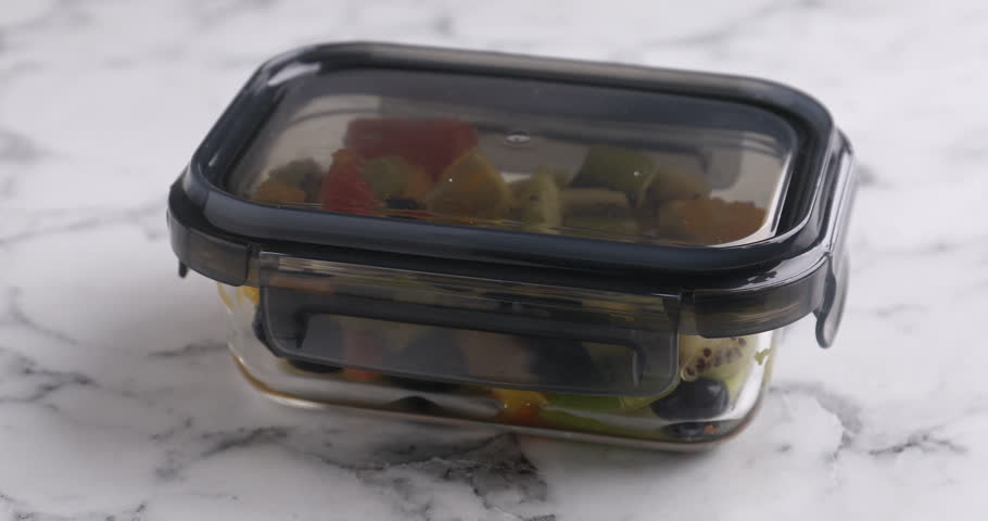 Woman opening glass container with tasty fruit salad at white marble table, closeup. Healthy snack