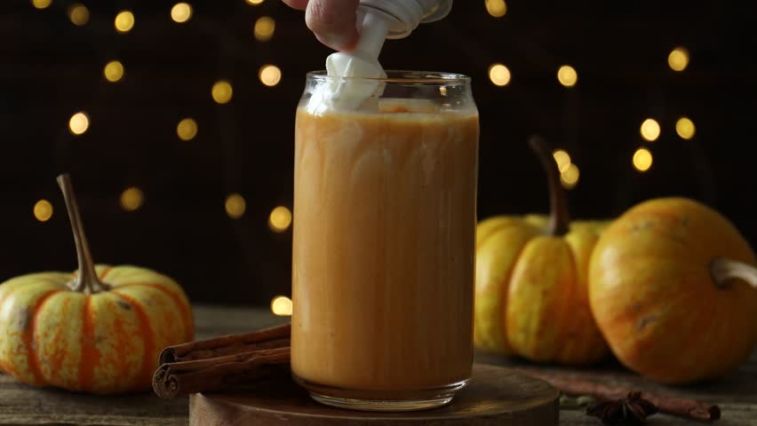 Woman adding whipped cream to tasty pumpkin latte at wooden table against blurred lights, closeup. Bokeh effect