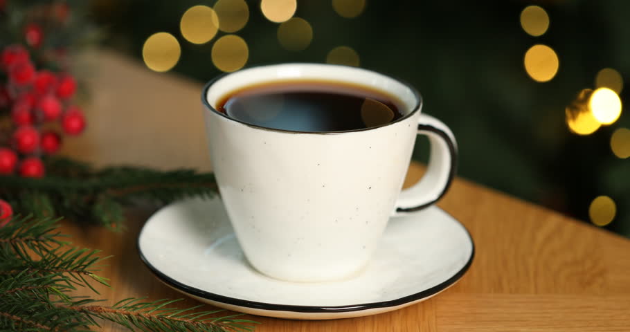 Aromatic coffee in cup and fir tree branches on wooden table against black background with flashing blurred lights, closeup