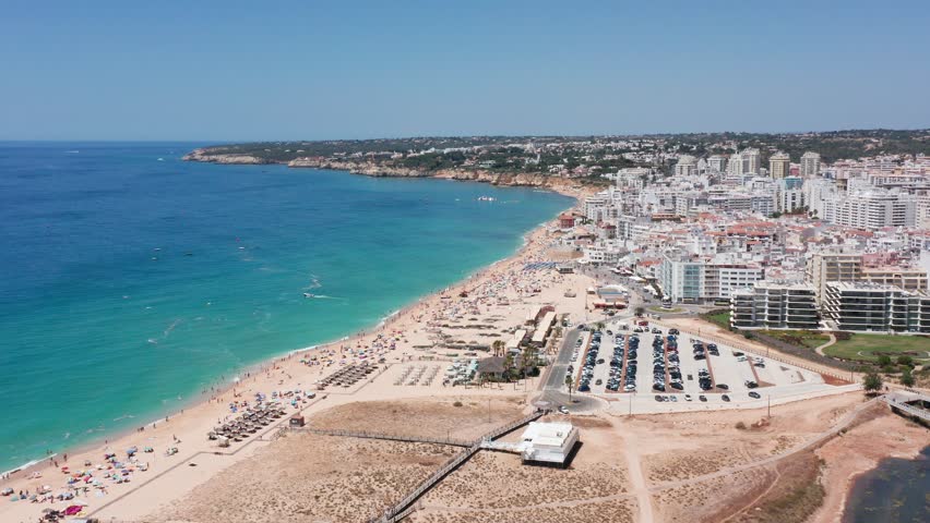 Seaside panorama view, Gentle waves hug shoreline near distant cityscape, Coastal landscape featuring sandy hills meeting clear blue lagoon with umbrellas and tranquil water