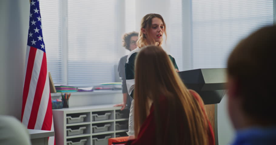American Female Student Delivers Speech With US Flag in Front of Classmates Sitting Desks. Concept Civic Education, Public Speaking Skills, and Student Voice in American Education System. Dolly Shot.