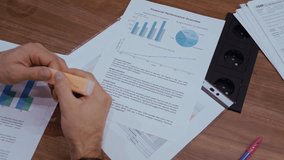 Detailed view of a business employee marking key points on a financial performance overview, emphasizing graphs and numerical data on a wooden table with scattered papers and visible outlets - Powered by Shutterstock - Get 15% off with code: PIKWIZARD15