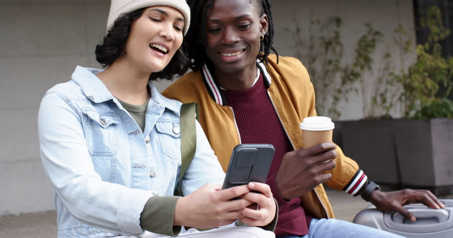 African American man holding coffee cup and woman on bench laughing at shared smartphone content. Casual, friendship, smiles, urban, communication, candid, lifestyle