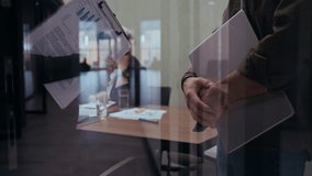 Two corporate professionals share a handshake after reaching agreement while studying data charts and financial documents. Shot through glass with reflection, colleague is seen behind them working. - Powered by Shutterstock - Get 15% off with code: PIKWIZARD15