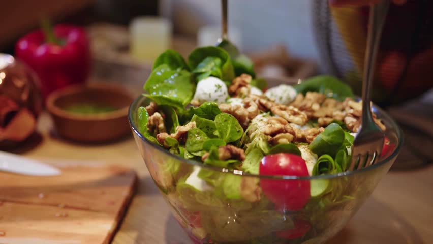 a close up shot of a salad being tossed. the salad contains walnuts, tomatoes, and cheese. it is in a glass bowl on a wooden table. a fork is mixing the salad. it looks delicious!