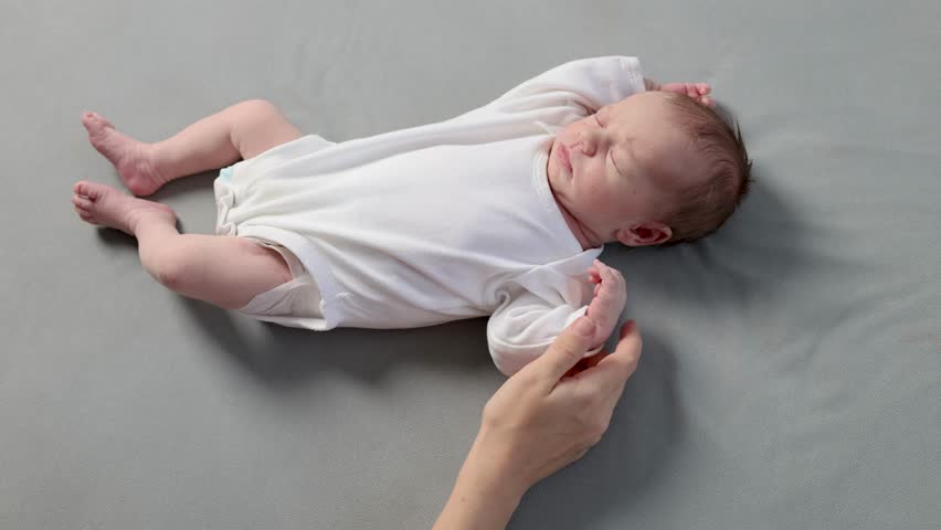 A newborn in a white bodysuit against a gray background. The little one is sleeping. The child is resting. The mother holds the baby