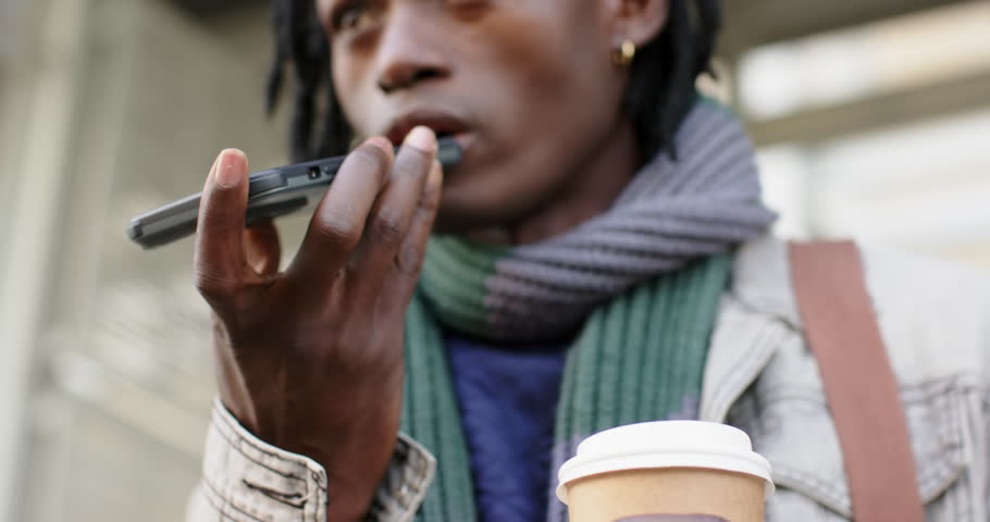 Mid adult Black man holding coffee cup raising smartphone recording message at bus shelter. Urban, communication, technology, realism, lifestyle, candid, street