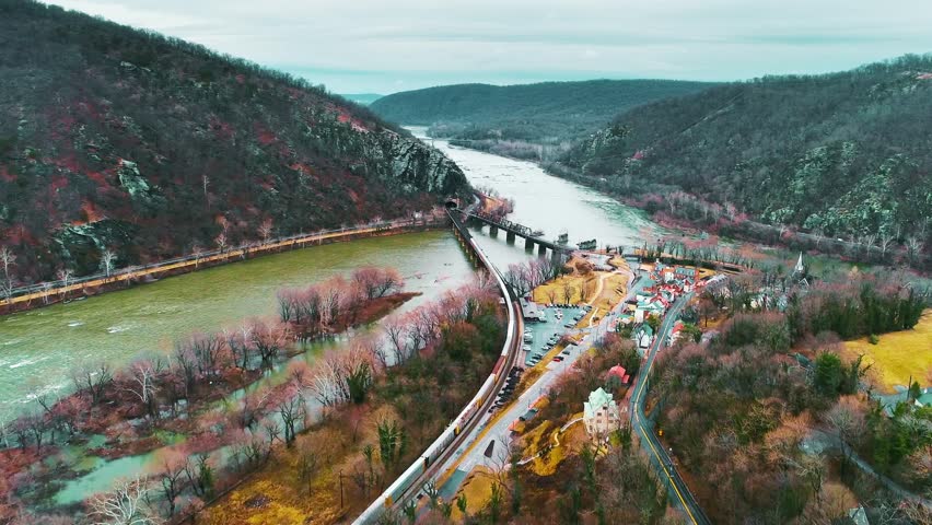 Cinematic aerial drone view of Harpers Ferry, West Virginia, showing the train bridge, Potomac River, and historic town surrounded by lush hills. Perfect for travel, history, and landscape visuals.